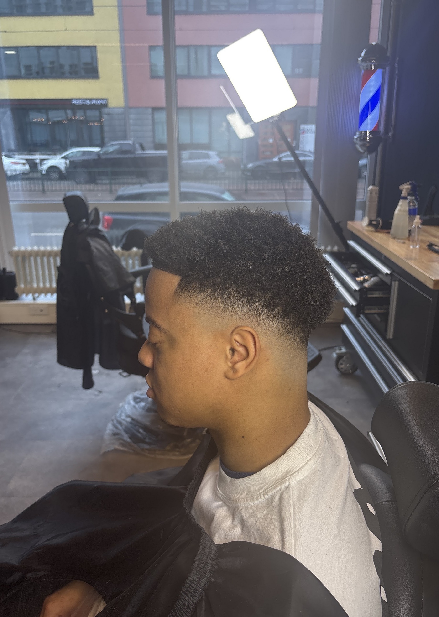Klant met een frisse fade, van opzij, bij het raam van de studio met de barberpaal erachter